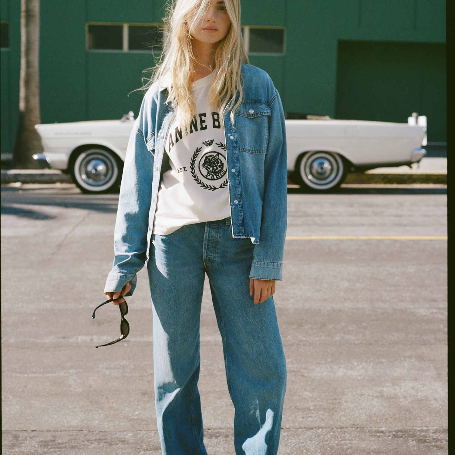 Person wearing a denim jacket and jeans standing in a parking lot with a vintage car in the background.