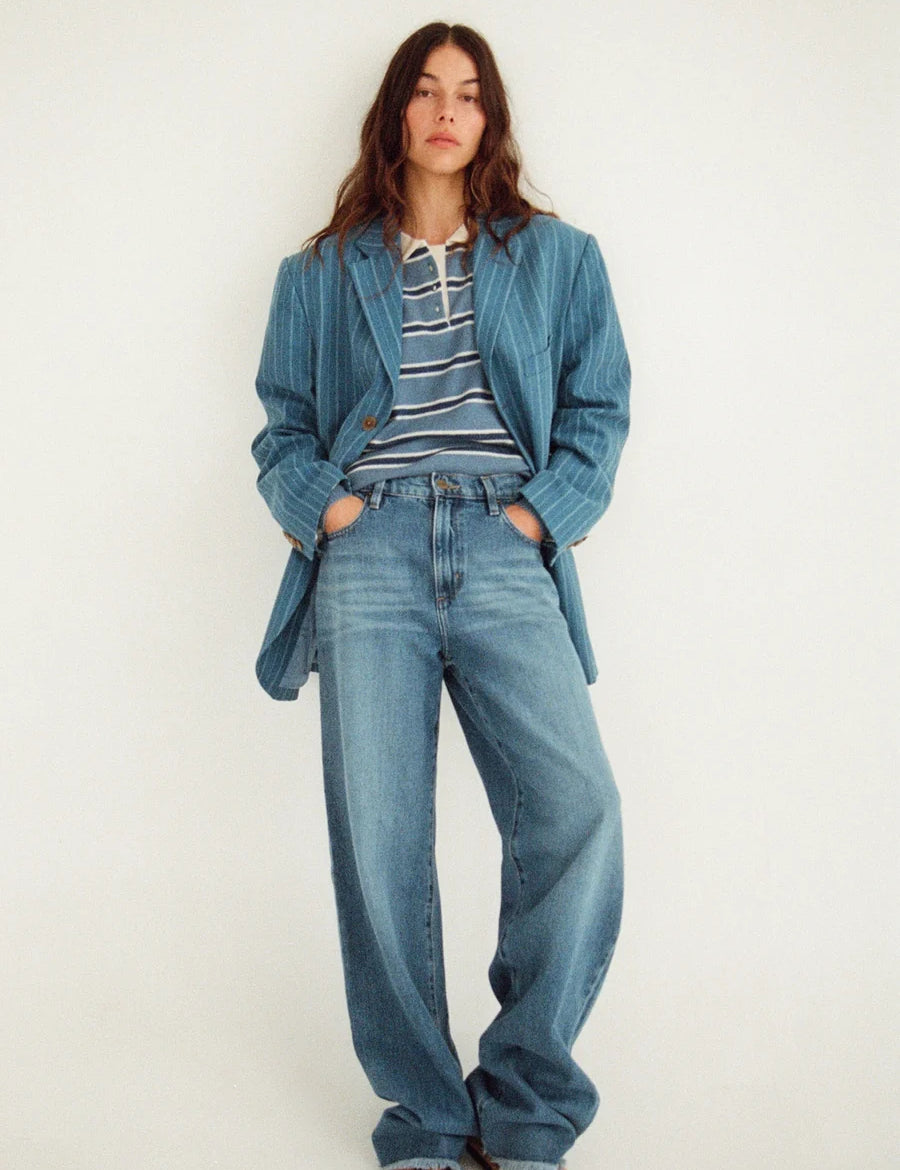 A woman stands against a plain light background wearing a blue corduroy blazer, wide-leg blue jeans, a navy and white striped t-shirt, and brown sandals.