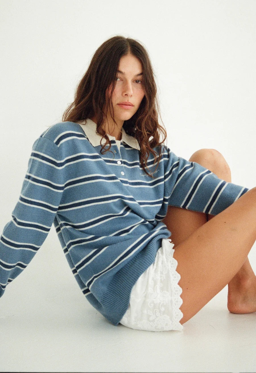 A woman sitting on a white studio floor wearing a blue and white striped long-sleeve knit polo shirt with a white collar, paired with a white skirt.