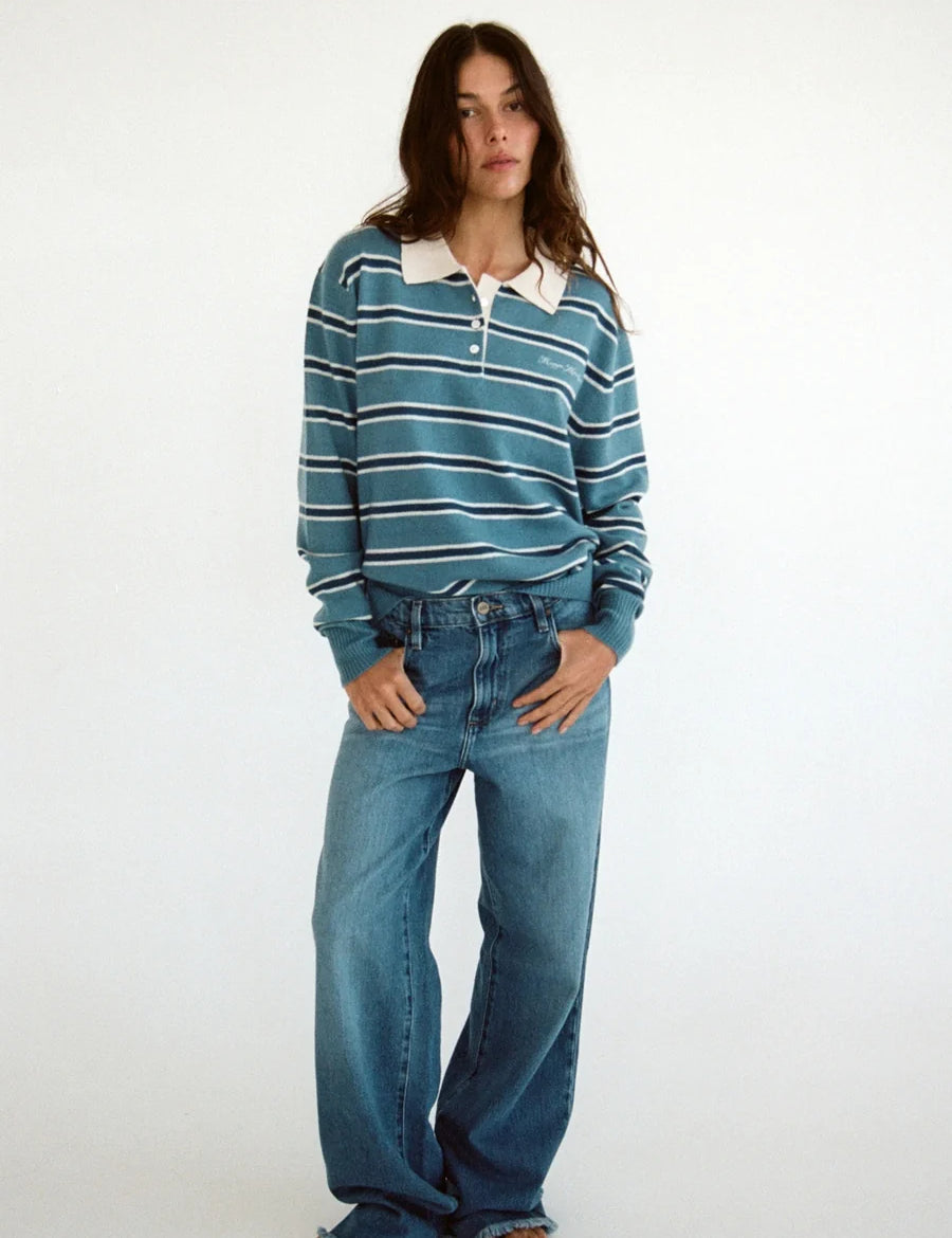 A woman modeling a teal and white striped long-sleeve polo shirt with blue wide-leg jeans and brown sandals, standing against a plain white studio background. cashmere 